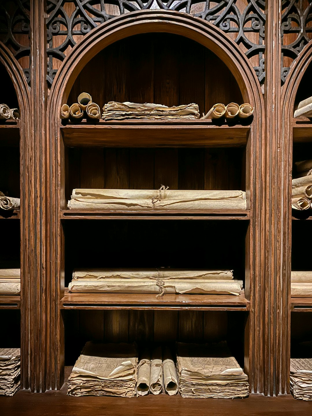 Pergamene antiche e carte fragili su scaffali di legno scuro in una biblioteca.